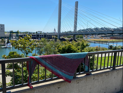 Knit shawl in blue and red draped over a railing with a bridge in the background