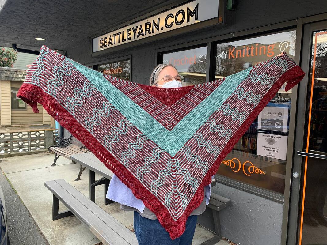 Power and Parallels shawl in blue and red held up in front of Seattle Yarn storefront